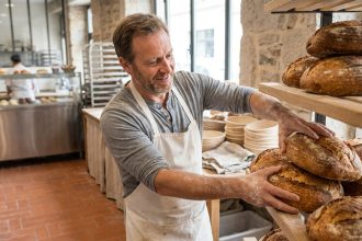 Boulanger professionnel organisant des pains artisanaux sur des étagères en bois dans une boulangerie bien rangée, séparée d'un espace pâtisserie en arrière-plan
